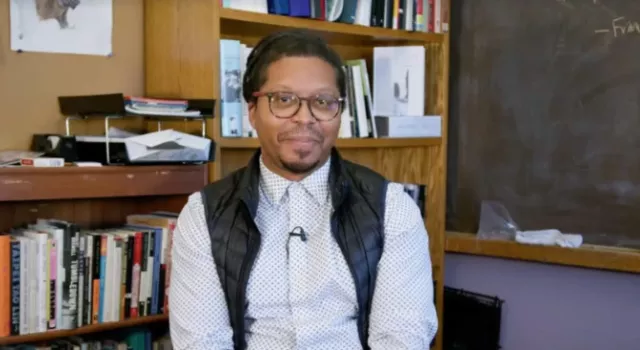 A person sitting in front of a bookshelf and chalkboard smiling.