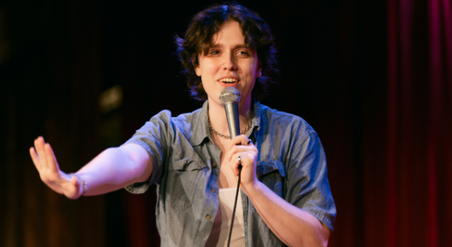 A person in a blue denim shirt over a white tank is standing on stage with a microphone in front of a red curtain smiling and holding out her hand.