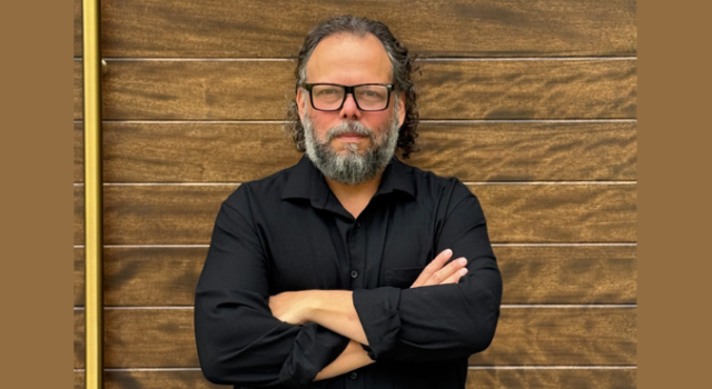 Person in glasses wearing a black button down shirt standing against a brown brick wall with arms crossed.