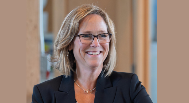 A person in a black blazer sitting in front of an office door and smiling. 