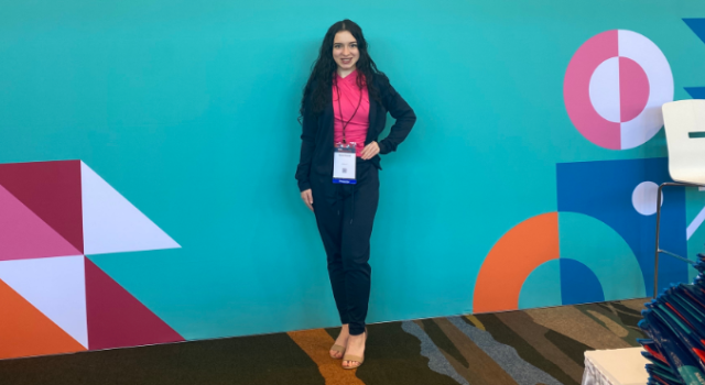 A person in a pink shirt and blue suit posing in front of a bright blue wall.