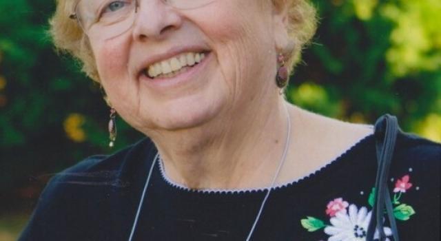 A person in glasses and a black top with flowers smiles standing outside in front of trees.