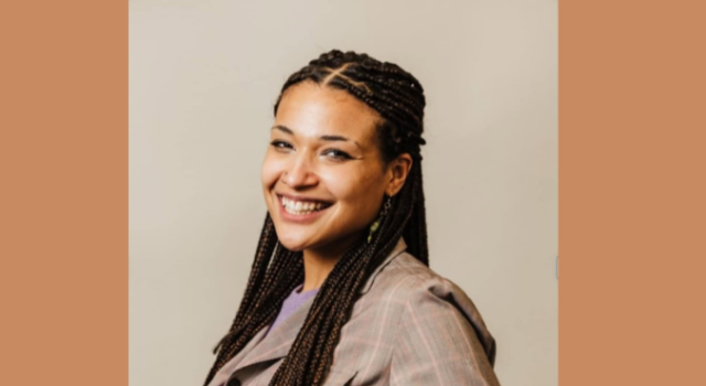 A woman turning toward the camera and smiling wearing a taupe shirt in front of a beige background.