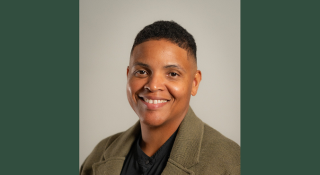 A person in a tan blazer and black shirt smiling against a plain background.