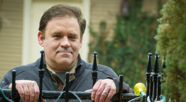 A person in a blue shirt stands behind a black iron fence and rests their hands on its top.