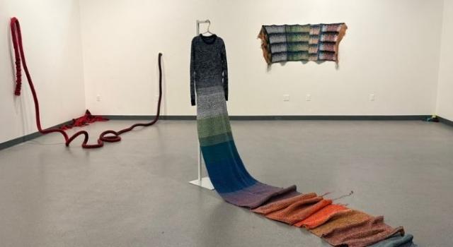 An art gallery featuring a colorful knitted dress with a long train on a dress form, a long, red knitted rope hanging off the wall and onto the floor, and a  colorful knit panel on the wall.
