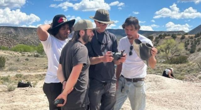 Four people standing in a huddle looking at a camera with a sweeping desert landscape behind them,