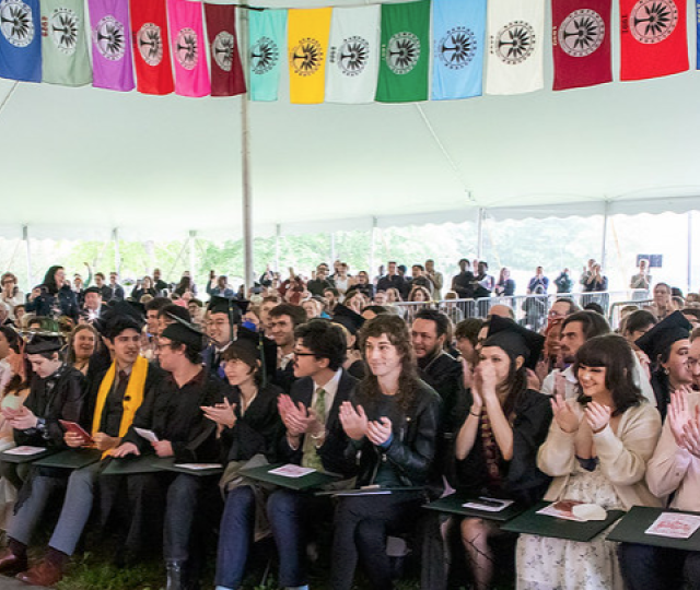 Graduates, faculty, staff members, and guests under the Commencement tent with colorful flags