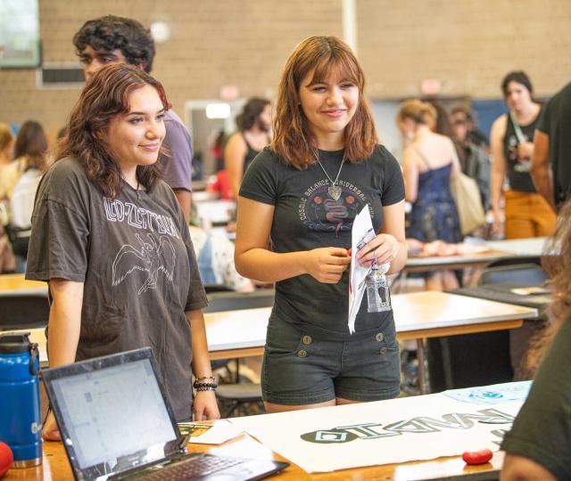 Two students are standing at a table at Hampfest learning more about an organization. They are both wearing tee shirts.