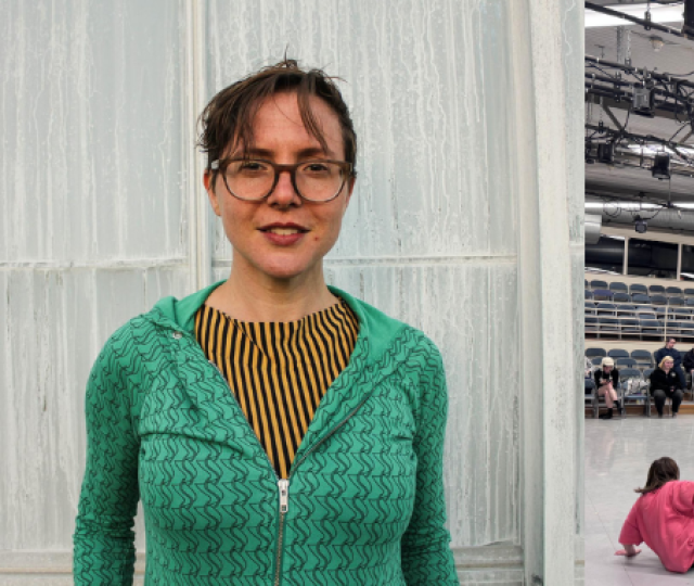 Split image showing a dance professor and a rehearsal space. On the left, a person with short brown hair, glasses, and a green zip-up sweater stands smiling in front of a frosted glass wall. On the right, dancers are spread across a large studio floor in various poses during a class or rehearsal, with an audience observing from bleachers in the background. The room is brightly lit with exposed rigging and stage lights overhead.