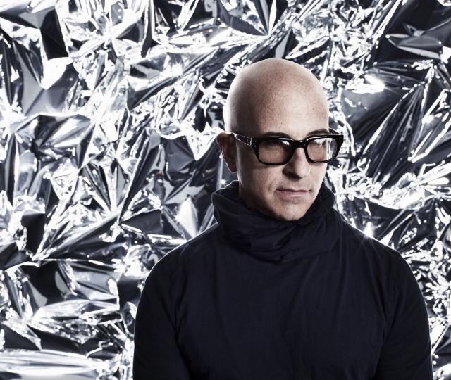 A person in glasses and a black turtleneck standing in front of a silver backdrop.