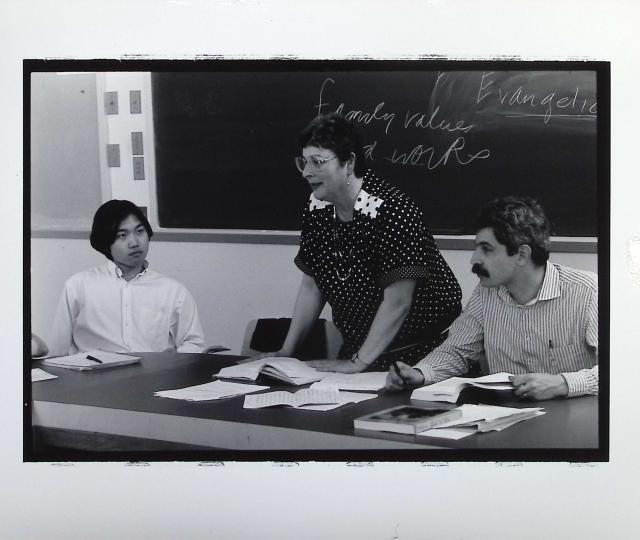 A person standing in front of a blackboard with people sitting to their right and left.