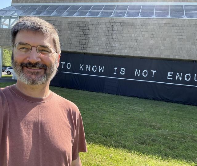 A person in glasses and a red shirt stands in front of a sign reading "to know is not enough."