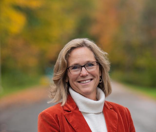 Jannifer Chrisler smiling, standing on a road with autumn foliage in the background