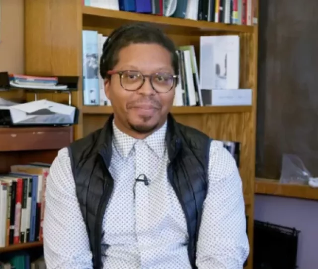 A person sitting in front of a bookshelf and chalkboard smiling.