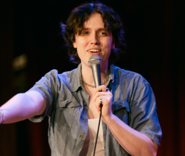 A person in a blue denim shirt over a white tank is standing on stage with a microphone in front of a red curtain smiling and holding out her hand.