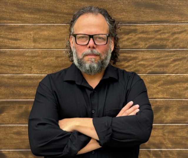 Person in glasses wearing a black button down shirt standing against a brown brick wall with arms crossed.
