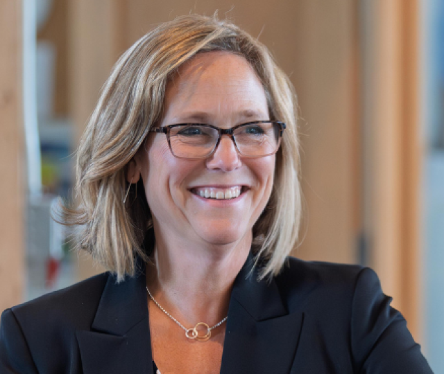 A person in a black blazer sitting in front of an office door and smiling. 