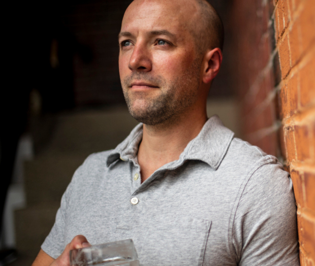 A person in a gray polo shirt standing against a brick wall holding a beer stein.