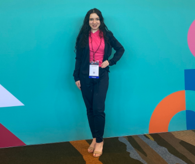 A person in a pink shirt and blue suit posing in front of a bright blue wall.