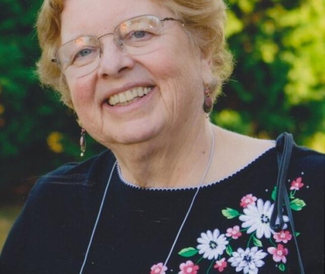 A person in glasses and a black top with flowers smiles standing outside in front of trees.