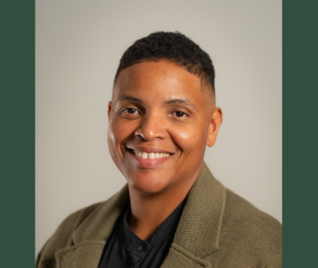 A person in a tan blazer and black shirt smiling against a plain background.
