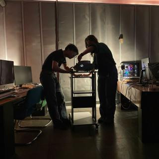 Alt text: Two people work together in a dimly lit room, adjusting a projector on a rolling cart. The projector casts a small glow, illuminating their hands. Surrounding them are desks with computer monitors, keyboards, and a custom-built PC with colorful LED lights. The walls are dark and industrial-looking, creating a moody atmosphere.