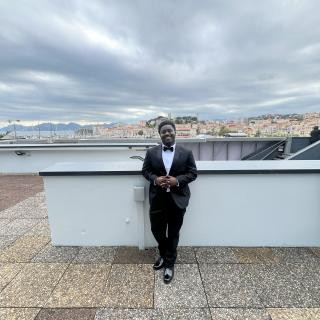 A person standing in a tuxedo in front of seaside buildings smiling.