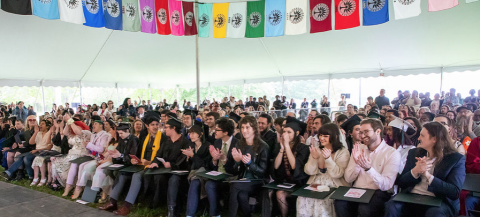Graduates, faculty, staff members, and guests under the Commencement tent with colorful flags