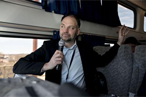 Stephen Gardner standing aboard an Amtrak train, holding a microphone in hand.