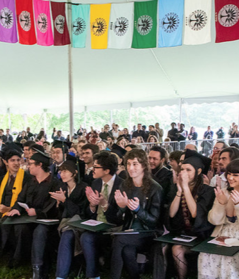 Graduates, faculty, staff members, and guests under the Commencement tent with colorful flags