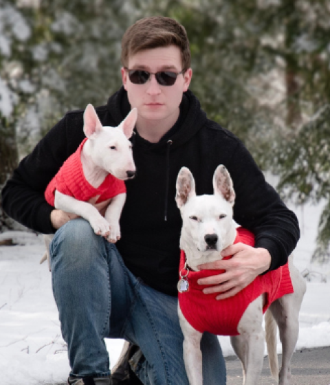 Dmitry Pepper crouching in a snowy landscape. He is holding two white dogs, both wearing red sweaters.