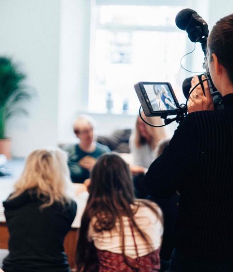 People are having a discussion while someone stands with a camera filming it.