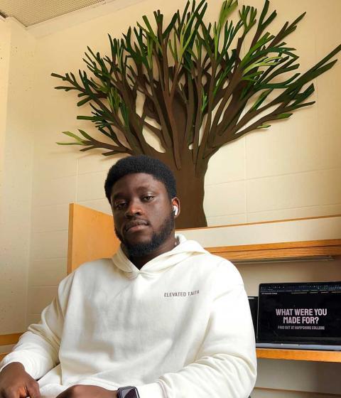 Rupert Tawiah-Quashie F22 wears a white sweatshirt and looks into the camera. He is sitting at a desk.