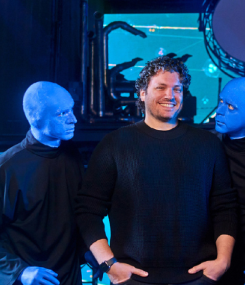 Two men stand in the center of a stage, surrounded by three performers from the Blue Man Group. The performers have their faces and hands painted blue and wear black clothing. One of them leans in toward the man on the left, who is smiling and wearing a black sweater, while the other two stand stoically. The man on the right, wearing a dark button-up shirt, crosses his arms with a slight smile. The background features a high-tech stage setup with LED screens displaying abstract visuals and circular lighting
