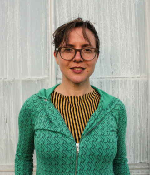 Split image showing a dance professor and a rehearsal space. On the left, a person with short brown hair, glasses, and a green zip-up sweater stands smiling in front of a frosted glass wall. On the right, dancers are spread across a large studio floor in various poses during a class or rehearsal, with an audience observing from bleachers in the background. The room is brightly lit with exposed rigging and stage lights overhead.