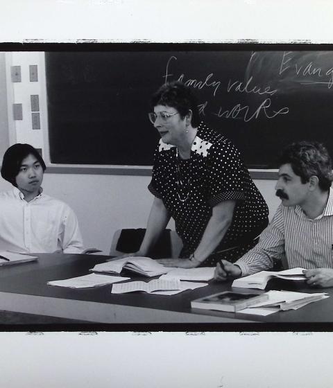 A person standing in front of a blackboard with people sitting to their right and left.