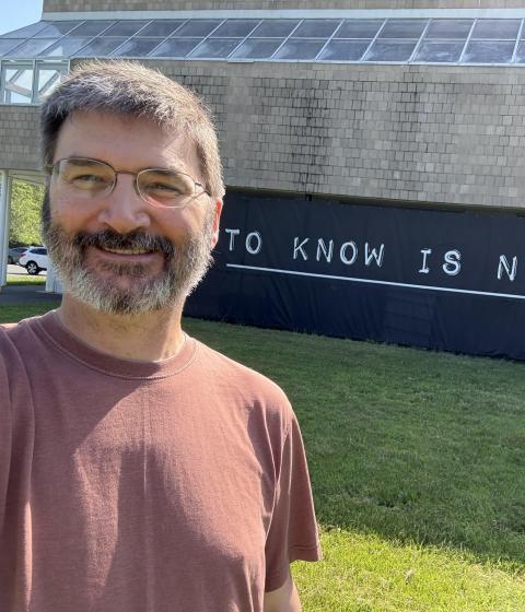 A person in glasses and a red shirt stands in front of a sign reading "to know is not enough."