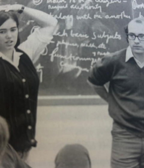 Two people stand in front of a blackboard with chalk writing on it.