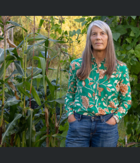 A person in a floral print shirt and jeans standing in front of a wall of plants.