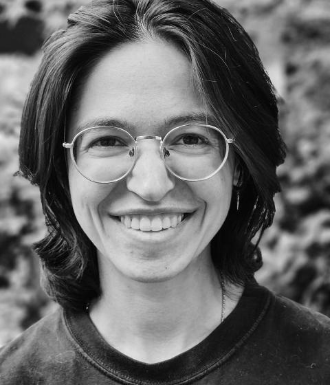 A black and white photo of a person standing in front of greenery and smiling.