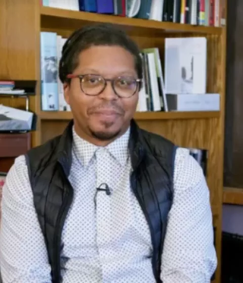A person sitting in front of a bookshelf and chalkboard smiling.