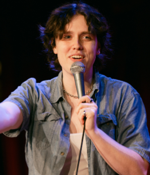A person in a blue denim shirt over a white tank is standing on stage with a microphone in front of a red curtain smiling and holding out her hand.