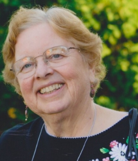 A person in glasses and a black top with flowers smiles standing outside in front of trees.