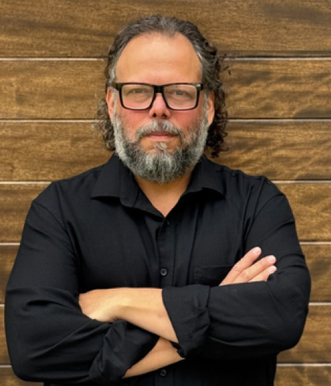 Person in glasses wearing a black button down shirt standing against a brown brick wall with arms crossed.