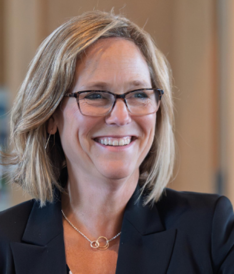 A person in a black blazer sitting in front of an office door and smiling. 