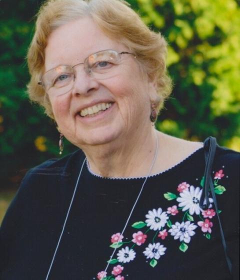 A person in glasses and a black top with flowers smiles standing outside in front of trees.