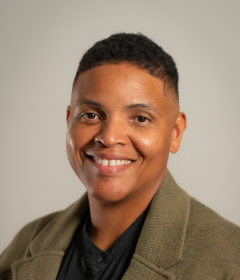 A person in a tan blazer and black shirt smiling against a plain background.