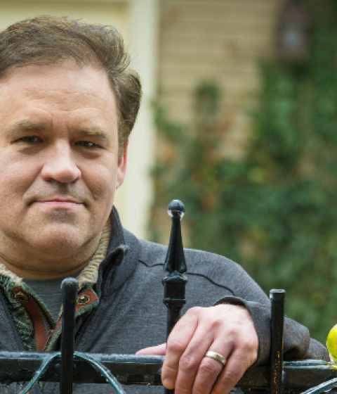A person in a blue shirt stands behind a black iron fence and rests their hands on its top.
