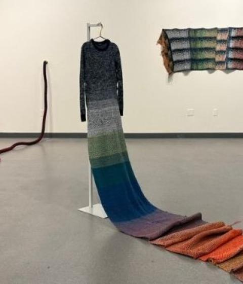 An art gallery featuring a colorful knitted dress with a long train on a dress form, a long, red knitted rope hanging off the wall and onto the floor, and a  colorful knit panel on the wall.